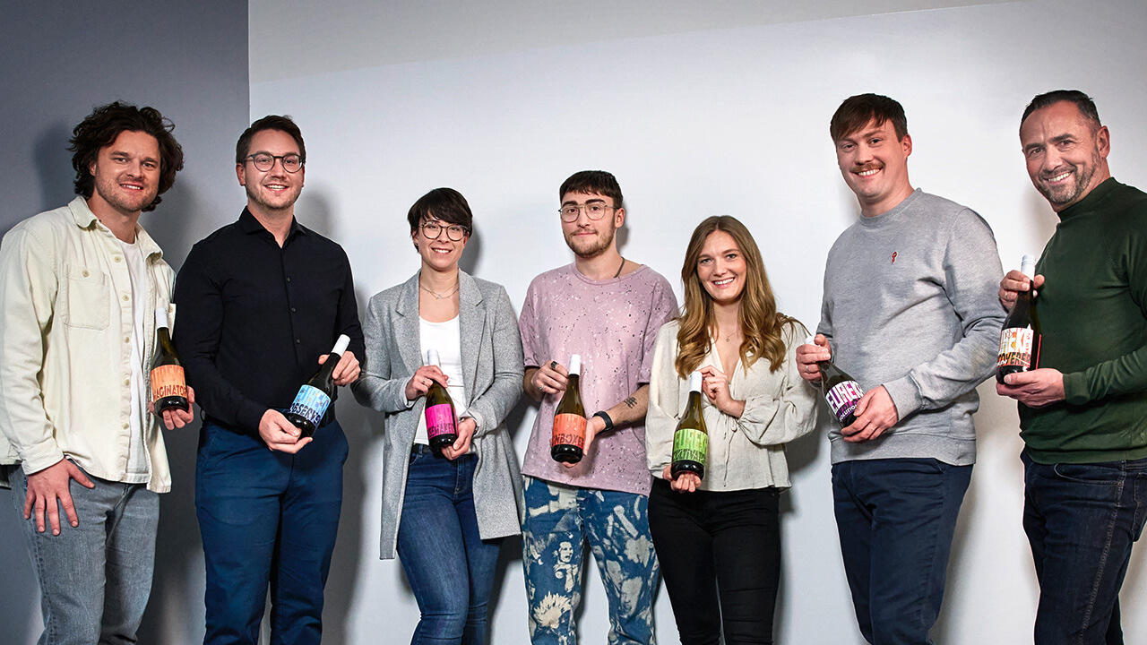 Sieben Menschen posieren jeweils mit einer Weinflasche mit Etikett in einer anderen Farbe
