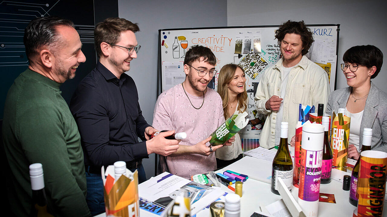 Six cheerful people gathered around a standing table examine bottle labels and packaging